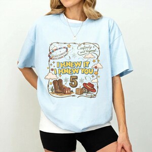 May include: A light blue t-shirt featuring a graphic print with the text "I KNEW IT I KNEW YOU", "5", and "Cinfinity and beyond". The design includes a lasso border, cowboy boots, a red hat, alphabet blocks, clouds, and stars.