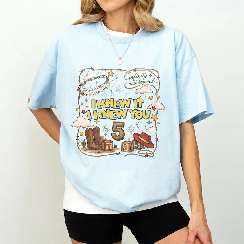 May include: A light blue t-shirt featuring a graphic print with the text "I KNEW IT I KNEW YOU", "5", and "Cinfinity and beyond". The design includes a lasso border, cowboy boots, a red hat, alphabet blocks, clouds, and stars.