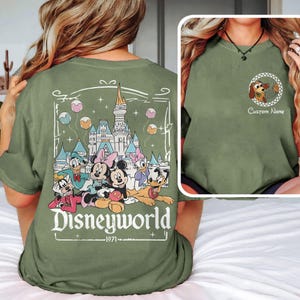 May include: Olive green t-shirt featuring a Disney World graphic on the back with Mickey, Minnie, Donald, Daisy, and Goofy. The front has a small circular graphic with a cartoon dog and the text "Custom Name".