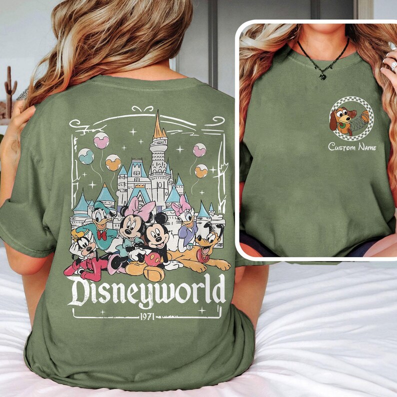 May include: Olive green t-shirt featuring a Disney World graphic on the back with Mickey, Minnie, Donald, Daisy, and Goofy. The front has a small circular graphic with a cartoon dog and the text "Custom Name".