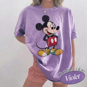 May include: A violet Comfort Colors t-shirt with a Mickey Mouse graphic. The cartoon character is in a classic pose, with red shorts, yellow shoes, and a black body. The shirt has a relaxed fit. The text 'Comfort Colors Violet' is visible.