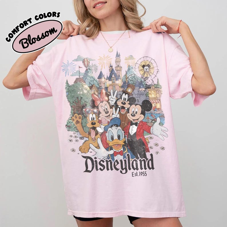 May include: A light pink t-shirt featuring a graphic print of Mickey Mouse, Minnie Mouse, Donald Duck, Goofy, and Pluto in front of a castle and fireworks. The text reads Disneyland Est. 1955. A Comfort Colors Blossom label is visible in the corner.