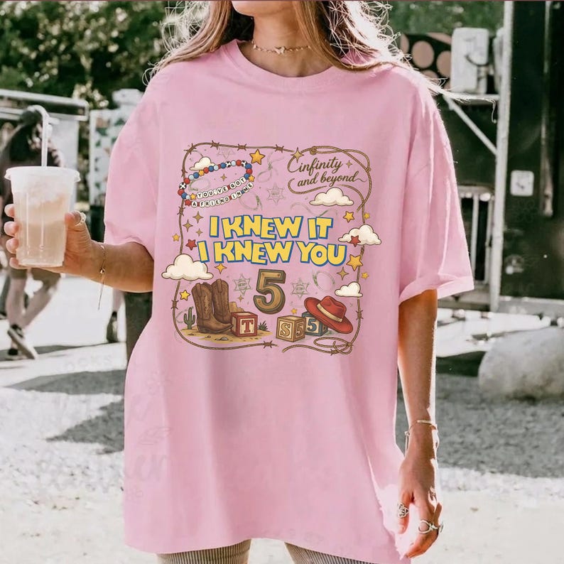 May include: A light pink t-shirt featuring a graphic print with the text "I KNEW IT I KNEW YOU", "5", and "Cinfinity and beyond". The design includes illustrations of cowboy boots, a cowboy hat, alphabet blocks, clouds, and a beaded bracelet.