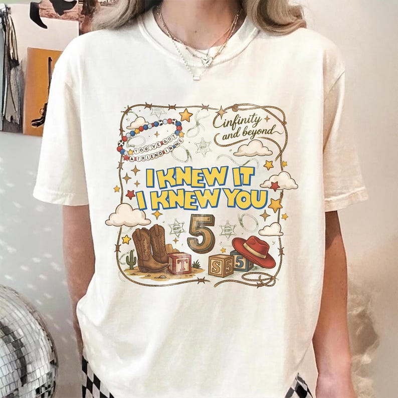 May include: A cream-colored short-sleeve t-shirt featuring a graphic print with the text "Cinfinity and beyond", "I KNEW IT", and "I KNEW YOU". The design includes a large number 5, cowboy boots, a red hat, alphabet blocks, and stars.