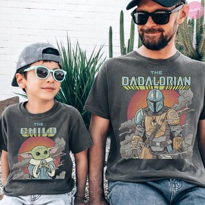 Może przedstawiać: Dwie pasujące antracytowe koszulki. Jedna koszulka zawiera napis "THE CHILD" z grafiką zielonego kosmity. Druga koszulka m&oacute;wi "THE DADALORIAN" z grafiką Mandalorianina.