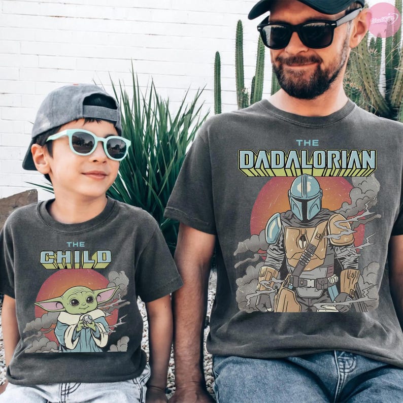 Może przedstawiać: Dwie pasujące antracytowe koszulki. Jedna koszulka zawiera napis "THE CHILD" z grafiką zielonego kosmity. Druga koszulka m&oacute;wi "THE DADALORIAN" z grafiką Mandalorianina.
