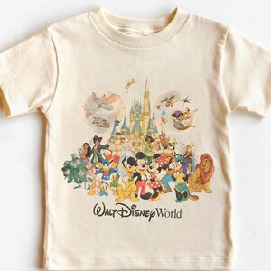 Puede incluir: Camiseta color crema con un gr&aacute;fico colorido de personajes de Disney, incluyendo Mickey y Minnie Mouse, Donald Duck y Simba. La imagen incluye un castillo y el texto "Walt Disney World".
