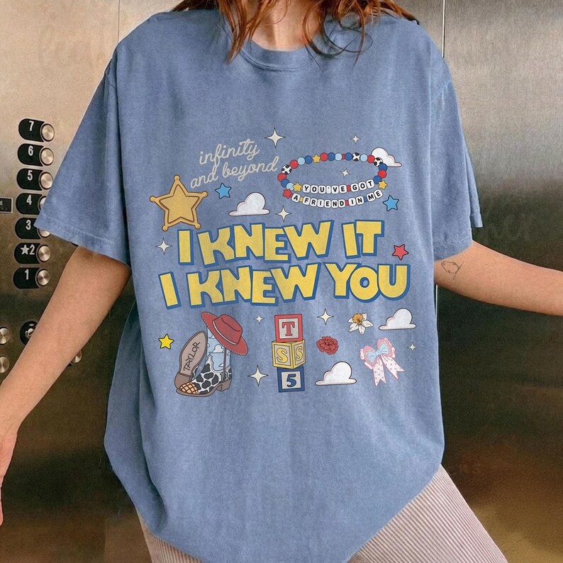 May include: A blue short-sleeve t-shirt featuring a graphic print with the text "infinity and beyond," "YOU'VE GOT A FRIEND IN ME," "I KNEW IT," and "I KNEW YOU." The design includes a yellow star, a beaded bracelet, a cowboy boot, and blocks.