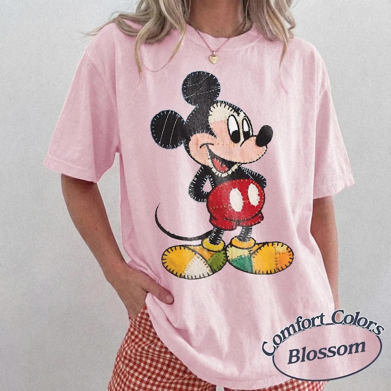 May include: A pale pink t-shirt with a stitched Mickey Mouse design. The cartoon character is depicted in a standing pose, wearing red shorts, yellow and green shoes, and black gloves. The shirt has the words "Comfort Colors Blossom" in a circular design.
