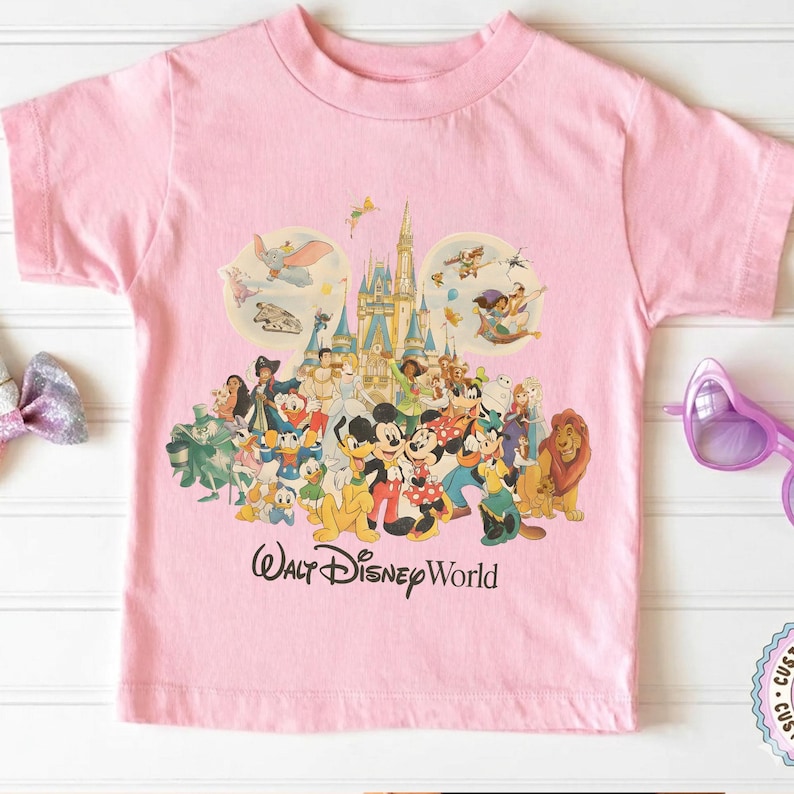 May include: Pink t-shirt featuring a colorful graphic of Disney characters, including Mickey and Minnie Mouse, and a castle. The words "Walt Disney World" are printed below the graphic. The shirt has short sleeves and a crew neck.