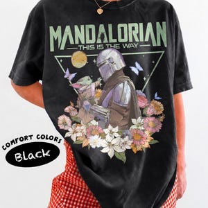 May include: Black t-shirt with a graphic of The Mandalorian and Grogu surrounded by flowers and butterflies. The text "MANDALORIAN - THIS IS THE WAY" is printed above the image. The shirt is labeled "COMFORT COLORS Black".