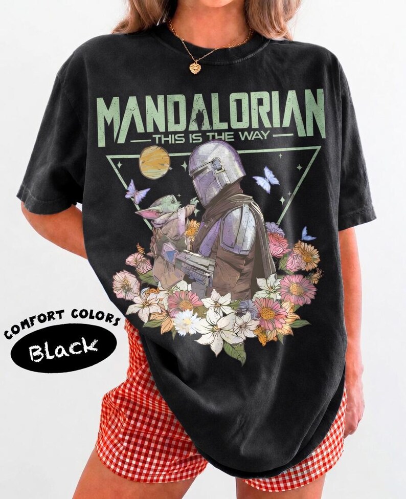 May include: Black t-shirt with a graphic of The Mandalorian and Grogu surrounded by flowers and butterflies. The text "MANDALORIAN - THIS IS THE WAY" is printed above the image. The shirt is labeled "COMFORT COLORS Black".