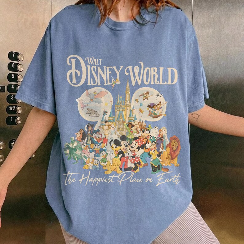 May include: Blue short-sleeved t-shirt featuring a Walt Disney World graphic. The design includes the park's logo, a castle, and various Disney characters. The text "The Happiest Place on Earth" is printed below the characters.