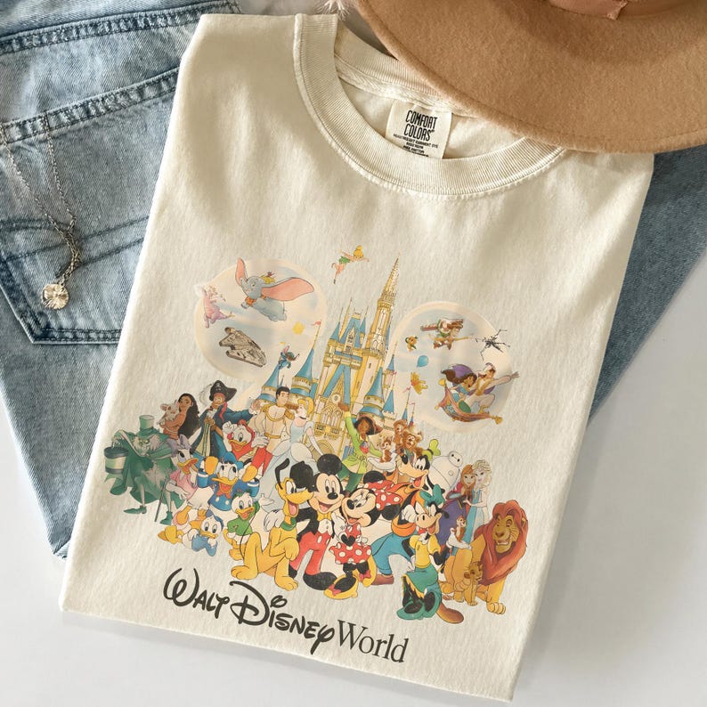 May include: Cream-colored t-shirt featuring a colorful graphic of Disney characters and Cinderella's castle. The words "Walt Disney World" are printed in black script at the bottom. The shirt is folded on a pair of blue jeans.