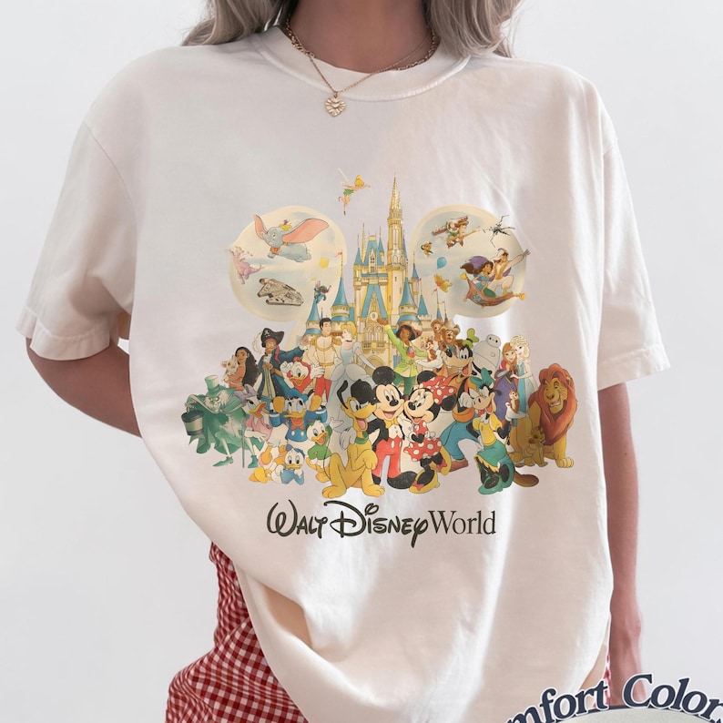May include: Cream-colored t-shirt featuring a colorful graphic of various Disney characters, including Mickey and Minnie Mouse, Donald Duck, and Simba. The words "Walt Disney World" are printed in a script font below the characters.