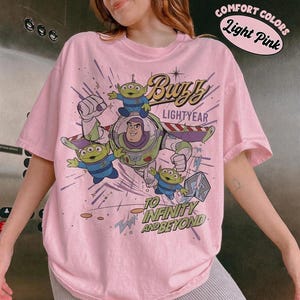 Pu&ograve; includere: T-shirt rosa chiaro con una grafica di Buzz Lightyear e gli alieni di Toy Story. Il design include il testo "Buzz Lightyear" e "To Infinity and Beyond". La maglietta ha anche il testo "Comfort Colors Light Pink".