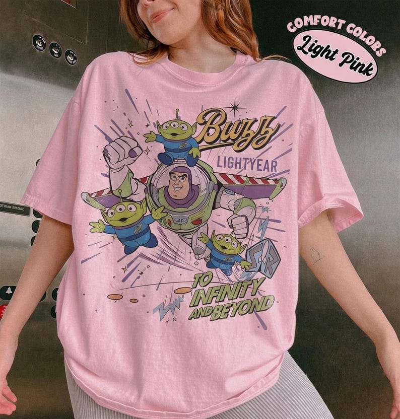 Pu&ograve; includere: T-shirt rosa chiaro con una grafica di Buzz Lightyear e gli alieni di Toy Story. Il design include il testo "Buzz Lightyear" e "To Infinity and Beyond". La maglietta ha anche il testo "Comfort Colors Light Pink".