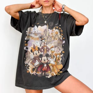 May include: A charcoal gray short-sleeve t-shirt featuring a graphic print of cartoon characters dressed in Halloween costumes. The design includes a castle, a ferris wheel, pumpkins, and a ghost, with characters wearing capes and hats.