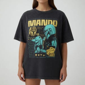 K&ouml;nnte beinhalten: Schwarzes T-Shirt mit einer Grafik von Mando und Grogu aus The Mandalorian. Das Design enth&auml;lt den Text "Mando" in Gelb und "Grogu" in T&uuml;rkis. Das T-Shirt hat einen Vintage-Look.