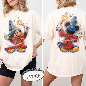 May include: An ivory t-shirt featuring a graphic of a cartoon mouse dressed as a wizard in a red robe and blue star-patterned hat. The front shows the mouse facing forward holding a glowing wand, while the back shows the mouse from behind.