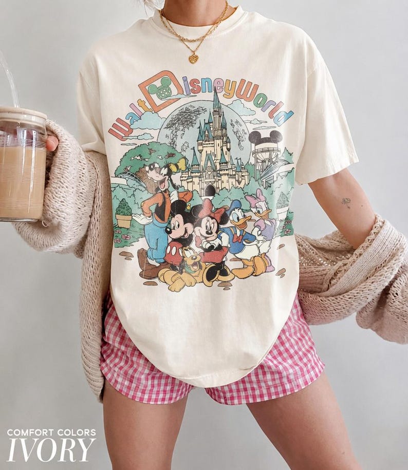 May include: Ivory-colored t-shirt featuring a colorful graphic of Disney characters and the text "Walt Disney World." The shirt is paired with pink and white checkered shorts. The words "Comfort Colors" and "Ivory" are printed at the bottom.