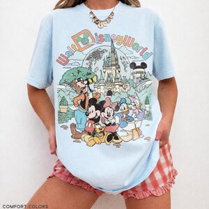 May include: Light blue t-shirt with a colorful Walt Disney World graphic featuring Mickey Mouse, Minnie Mouse, Donald Duck, and other characters. The shirt has the text "Walt Disney World" in a retro font. The bottom of the shirt has the text "COMFORT COLORS CHAMBRAY".