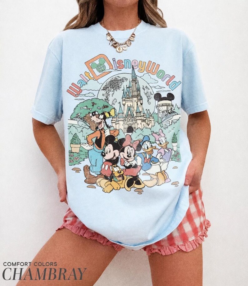 May include: Light blue t-shirt with a colorful Walt Disney World graphic featuring Mickey Mouse, Minnie Mouse, Donald Duck, and other characters. The shirt has the text "Walt Disney World" in a retro font. The bottom of the shirt has the text "COMFORT COLORS CHAMBRAY".
