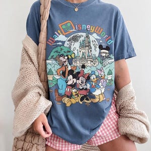 May include: Blue jean colored t-shirt featuring a colorful graphic of Disney World characters and the park's logo. The shirt has a vintage look and feel. The words "COMFORT COLORS" and "BLUE JEAN" are printed at the bottom of the image.