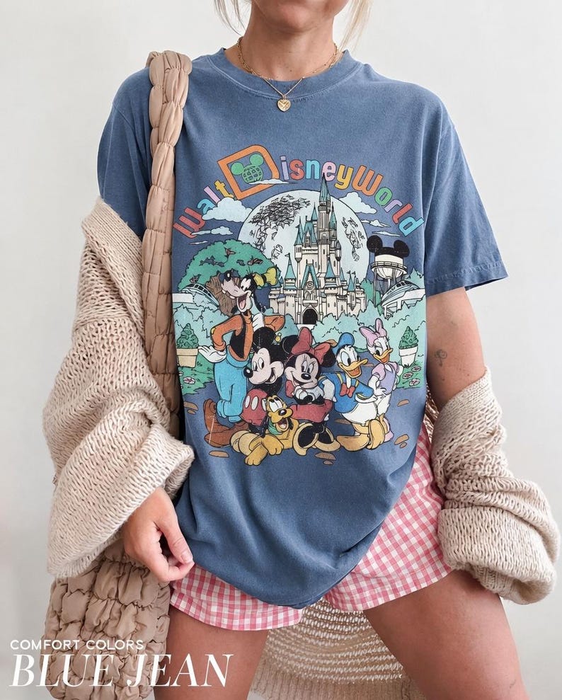 May include: Blue jean colored t-shirt featuring a colorful graphic of Disney World characters and the park's logo. The shirt has a vintage look and feel. The words "COMFORT COLORS" and "BLUE JEAN" are printed at the bottom of the image.