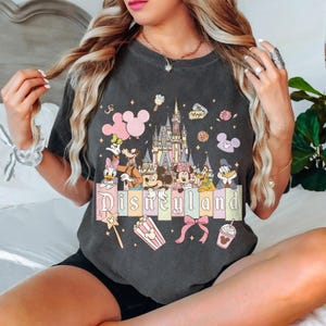 May include: Dark gray t-shirt featuring a colorful Disneyland graphic. The design includes Disney characters, a castle, and the word "Disneyland" in pastel colors. Additional elements include Mickey Mouse icons, popcorn, and a bow.