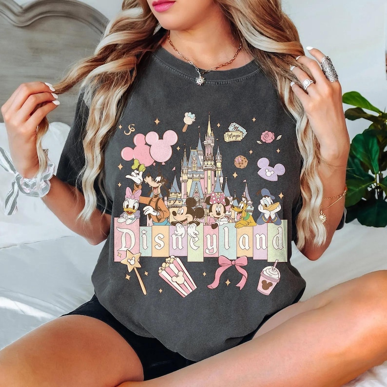 May include: Dark gray t-shirt featuring a colorful Disneyland graphic. The design includes Disney characters, a castle, and the word "Disneyland" in pastel colors. Additional elements include Mickey Mouse icons, popcorn, and a bow.