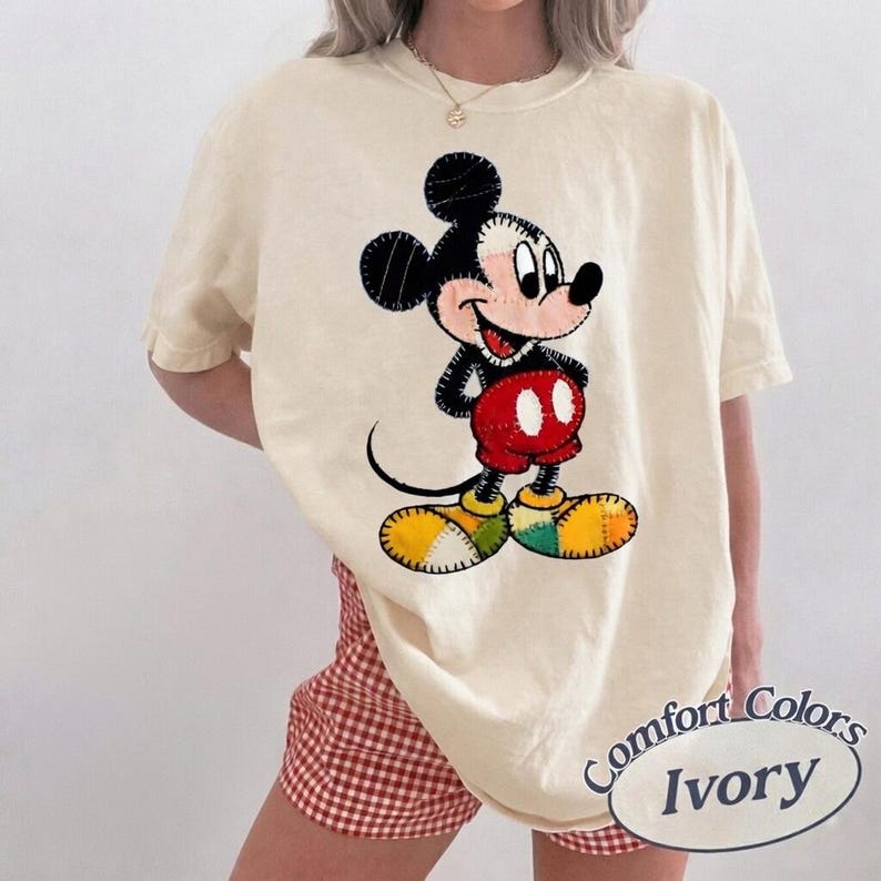 May include: Ivory-coloured t-shirt with a colourful Mickey Mouse graphic. The cartoon character is depicted in black, red, yellow, and green patches. The shirt has short sleeves and a relaxed fit. The text "Comfort Colors Ivory" is printed on the bottom right.