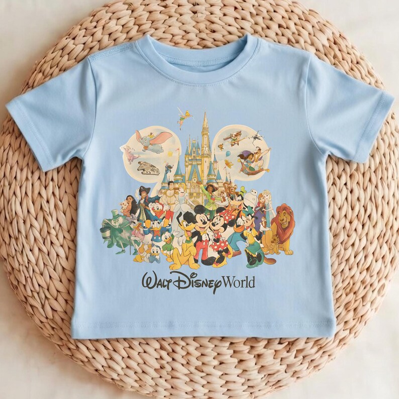 Puede incluir: Una camiseta azul claro con un gr&aacute;fico colorido de varios personajes de Disney, incluyendo Mickey Mouse, Donald Duck y Simba, frente a un castillo. Las palabras "Walt Disney World" est&aacute;n impresas debajo.