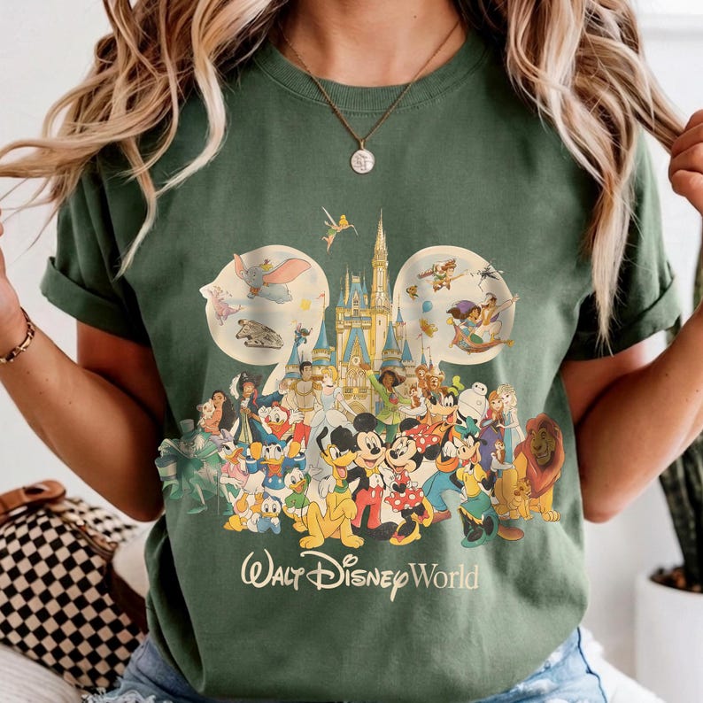 May include: Green t-shirt featuring a colorful graphic of various Disney characters, including Mickey Mouse, Donald Duck, and Simba. The design includes the text "Walt Disney World" in a stylized font. The shirt has short sleeves.