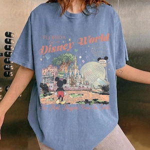 Puede incluir: Una camiseta azul con un gr&aacute;fico colorido de Disney World en Florida. El dise&ntilde;o incluye el texto "Florida Disney World" e im&aacute;genes de la Hollywood Tower, el castillo de Cenicienta y Spaceship Earth. Mickey Mouse tambi&eacute;n est&aacute; presente.