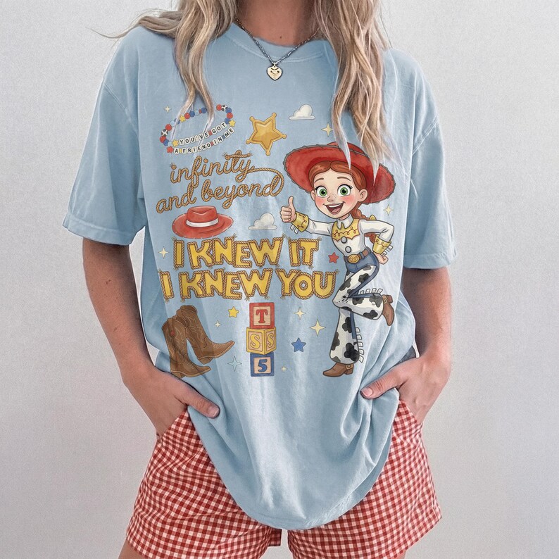 May include: A light blue short-sleeve t-shirt featuring a graphic print of a cowgirl character, a red hat, brown boots, and alphabet blocks. The text reads "infinity and beyond" and "I KNEW IT I KNEW YOU" along with "YOU'VE GOT A FRIEND IN ME".