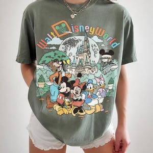 May include: Olive green t-shirt featuring a colorful graphic of Disney characters, including Mickey Mouse, Minnie Mouse, Donald Duck, and Goofy, in front of Cinderella's castle. The text "Walt Disney World" is arched across the top.