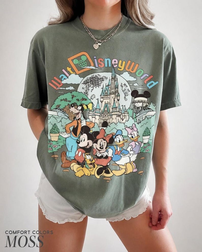 May include: Olive green t-shirt featuring a colorful graphic of Disney characters, including Mickey Mouse, Minnie Mouse, Donald Duck, and Goofy, in front of Cinderella's castle. The text "Walt Disney World" is arched across the top.