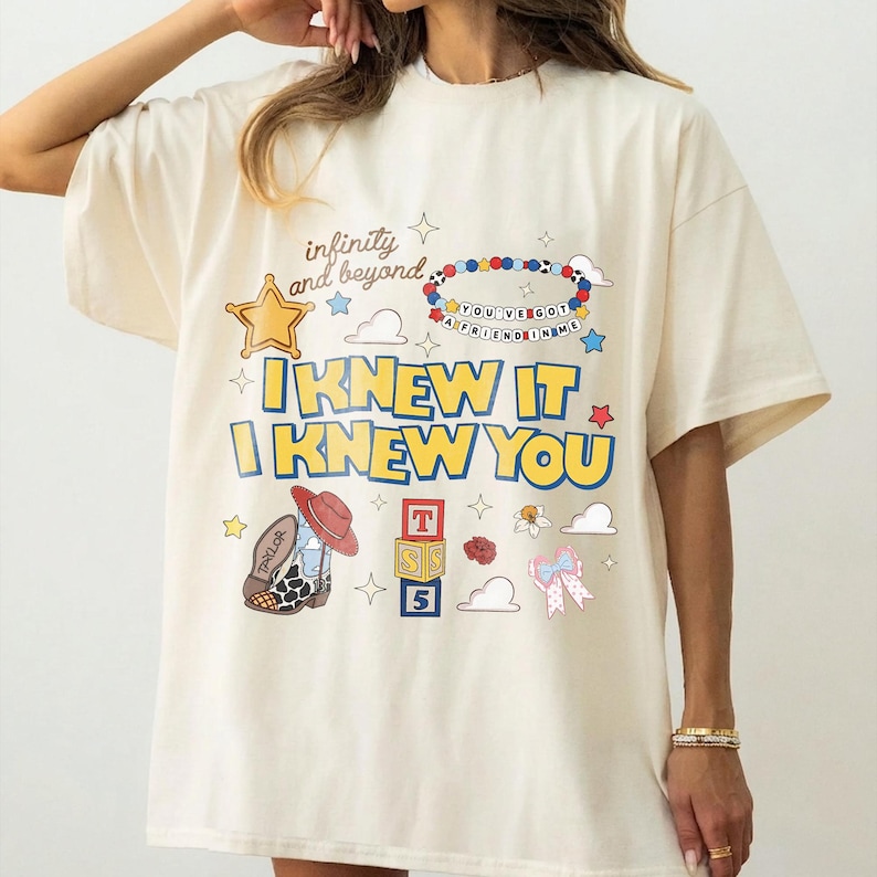 May include: An off-white t-shirt featuring a graphic print with the text "infinity and beyond," "YOU'VE GOT A FRIEND IN ME," "I KNEW IT," and "I KNEW YOU." The design includes a yellow star, a beaded bracelet, a cowboy boot, alphabet blocks, and a bow.