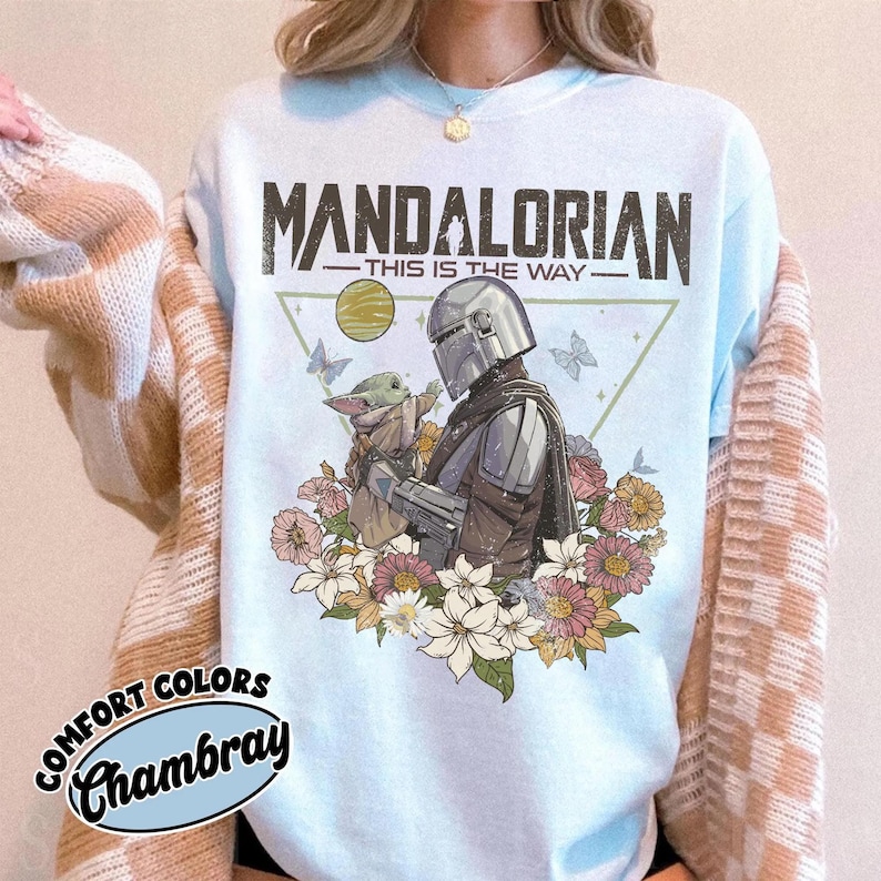 May include: Light blue t-shirt featuring a graphic of The Mandalorian and Grogu surrounded by flowers and butterflies. The text "MANDALORIAN - THIS IS THE WAY" is printed across the top. The shirt has the words "COMFORT COLORS Chambray" printed on the bottom left.