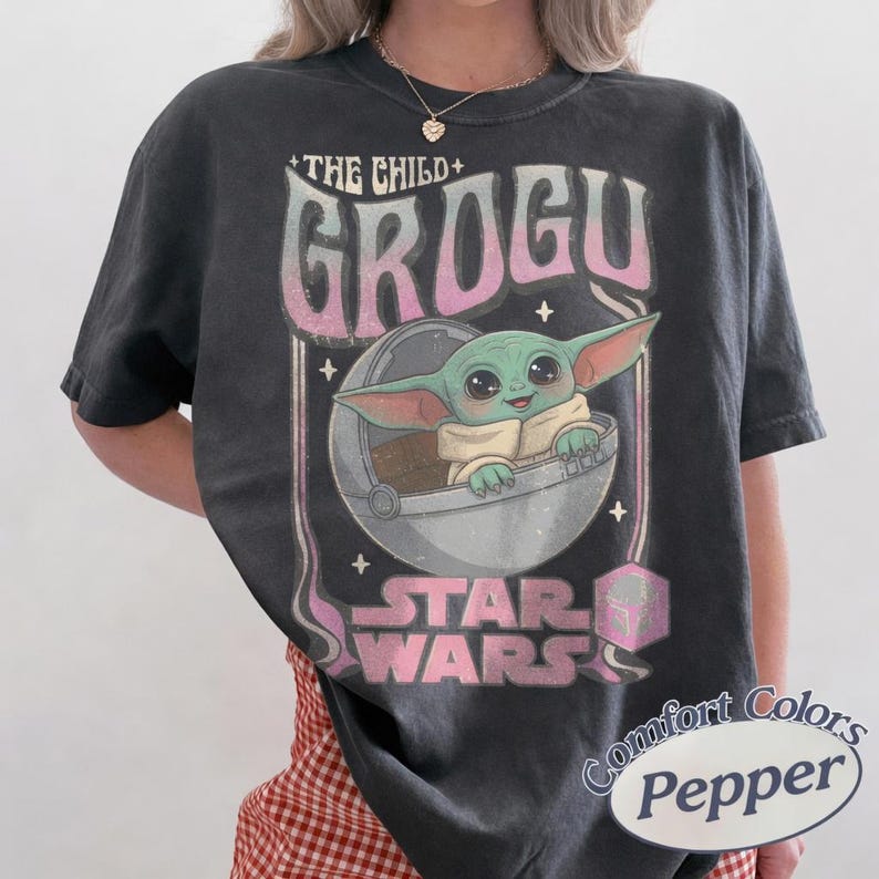 Puede incluir: Camiseta gris oscuro con Grogu de Star Wars. El dise&ntilde;o incluye el texto "THE CHILD + GROGU" en una fuente retro, con Grogu en su c&aacute;psula. El logotipo de Star Wars tambi&eacute;n es visible.