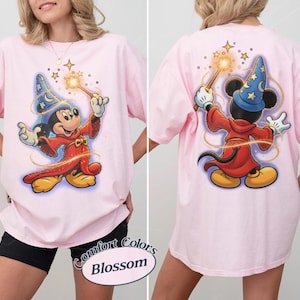 May include: A light pink t-shirt featuring a graphic of Mickey Mouse as a sorcerer. The front shows Mickey facing forward with a wand, while the back shows Mickey from behind casting a spell. The text Comfort Colors Blossom is printed below.