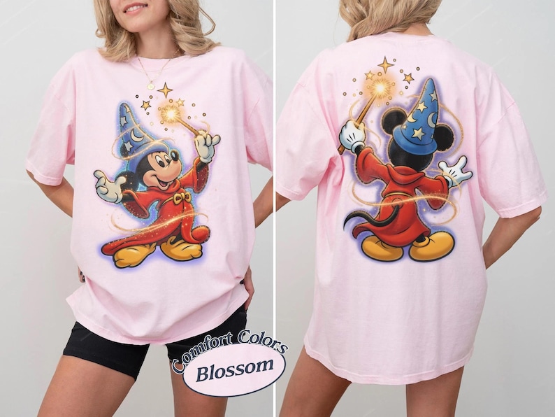 May include: A light pink t-shirt featuring a graphic of Mickey Mouse as a sorcerer. The front shows Mickey facing forward with a wand, while the back shows Mickey from behind casting a spell. The text Comfort Colors Blossom is printed below.