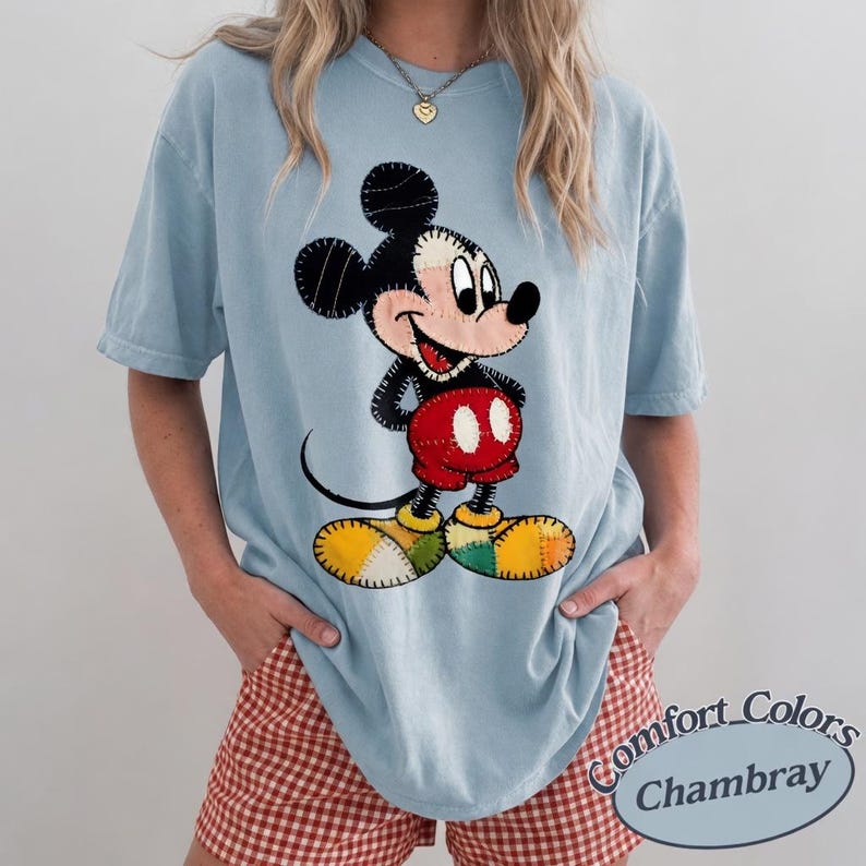 May include: Pale blue Comfort Colors Chambray t-shirt with a Mickey Mouse graphic. The graphic is a patchwork design in black, red, yellow, and green. The shirt has short sleeves and a loose fit. The text "Comfort Colors Chambray" is visible.