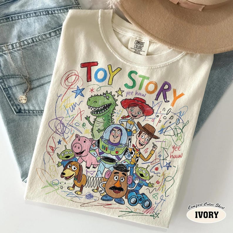 May include: Ivory-colored Comfort Colors t-shirt featuring a colorful "Toy Story" graphic. The design includes characters like Woody, Buzz Lightyear, and Rex, with the words "Toy Story" in a playful font. The shirt is laid on a pair of blue jeans.
