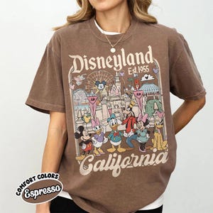 Peut inclure: T-shirt marron avec un motif Disneyland. Le design comprend Mickey Mouse, Minnie Mouse, Donald Duck, Dingo et Pluto devant une sc&egrave;ne de parc. Le texte "Disneyland Est. 1955 California" est &eacute;galement affich&eacute;.