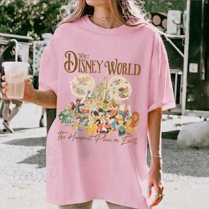 May include: Pink t-shirt featuring a Walt Disney World graphic. The design includes the words "Walt Disney World" in gold script above an image of Cinderella's castle and various Disney characters. The text "The Happiest Place on Earth" is below.