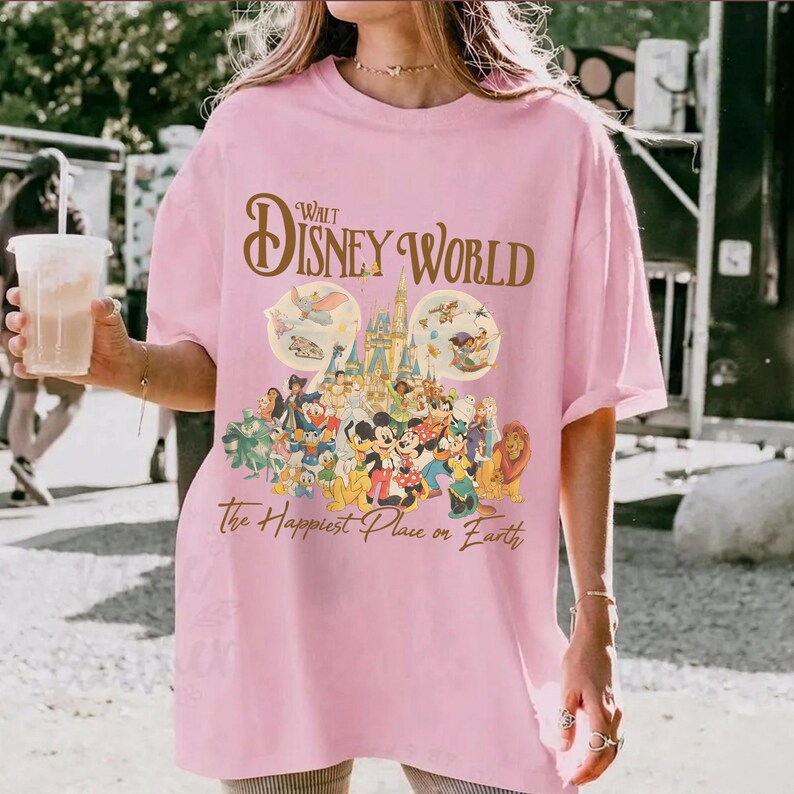 May include: Pink t-shirt featuring a Walt Disney World graphic. The design includes the words "Walt Disney World" in gold script above an image of Cinderella's castle and various Disney characters. The text "The Happiest Place on Earth" is below.