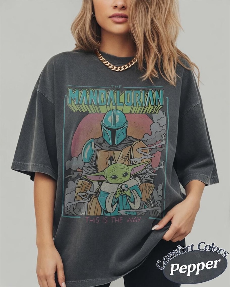May include: Dark grey t-shirt featuring a graphic of The Mandalorian and Grogu, with the text "THE MANDALORIAN" and "THIS IS THE WAY". The design includes a helmeted figure and a green alien. The shirt is labeled "Comfort Colors Pepper".