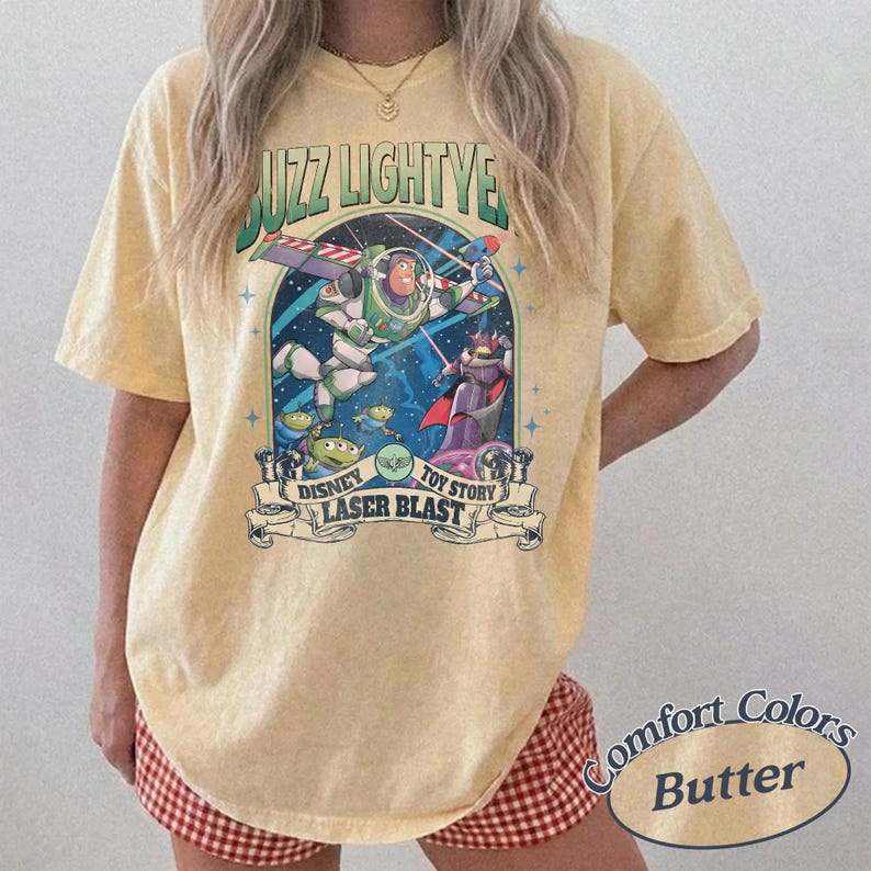 May include: A pale yellow short-sleeve t-shirt featuring a graphic print of Buzz Lightyear and other characters in space. The text reads BUZZ LIGHTYEAR, DISNEY TOY STORY, and LASER BLAST. The shirt has a crew neckline and a relaxed, loose fit.
